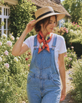 A woman standing in a sunlit garden wearing light blue denim overalls over a white T-shirt, with a bright orange floral neck scarf and a straw hat with a black band.