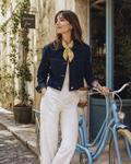 A woman stands on a cobblestone street next to a light blue bicycle, wearing a dark navy cropped denim jacket, white wide-leg trousers, and a yellow patterned silk neck scarf. She is posing in front of a rustic stone building with a hanging 'gocceria fine' sign.