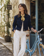 A woman stands on a cobblestone street next to a light blue bicycle, wearing a dark navy cropped denim jacket, white wide-leg trousers, and a yellow patterned silk neck scarf. She is posing in front of a rustic stone building with a hanging 'gocceria fine' sign.