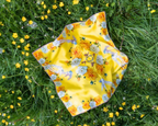 A square yellow scarf with a floral and bird pattern, laid flat on a green grassy field with small yellow flowers.