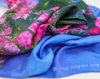 A close-up of draped fabric showing two colorways: a solid bright blue fabric with white vertical text reading 'THE PEONY GIRL' and a green floral print with large pink flowers.