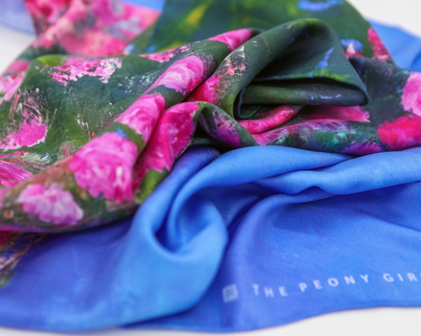 A close-up of draped fabric showing two colorways: a solid bright blue fabric with white vertical text reading 'THE PEONY GIRL' and a green floral print with large pink flowers.