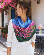 A woman wearing a white blouse and white pants models a square scarf with a vibrant floral print. The scarf features a blue sky-like top section and a lower section of pink flowers with green leaves, set against a sunny outdoor patio background.