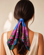 A woman shown from the back wearing a cream-colored sleeveless top, with her dark brown hair tied back in a ponytail. A vibrant silk scarf with a pink and purple floral pattern and blue borders is tied around her ponytail.