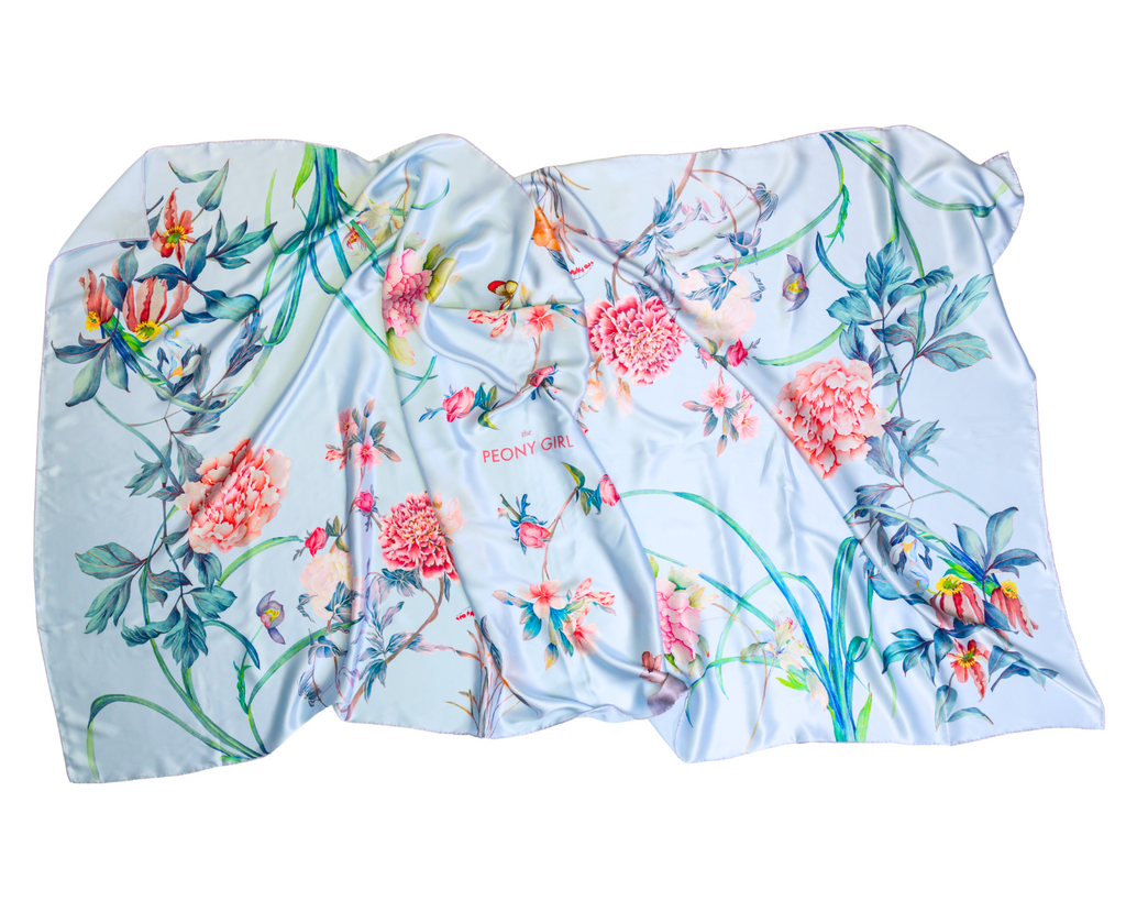 A light blue silk scarf with a vibrant watercolor floral print featuring pink peonies, green leaves, and small birds, laid flat on a white background.