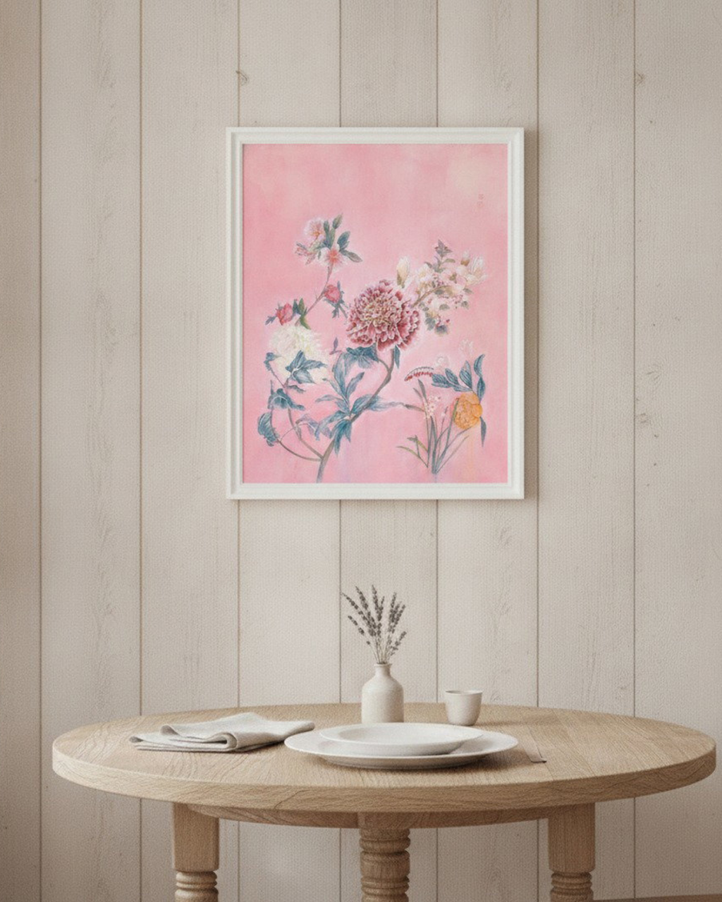 A framed floral art print with a pink background and botanical illustration, hanging on a light wood-paneled wall above a round wooden table with plates and a small vase.