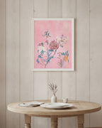 A framed floral art print with a pink background and botanical illustration, hanging on a light wood-paneled wall above a round wooden table with plates and a small vase.