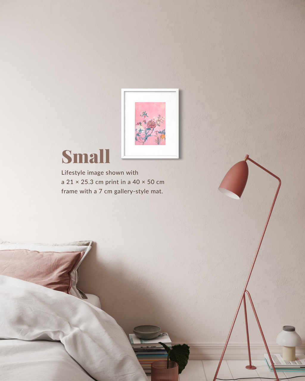 A framed art print with a pink floral design is displayed on a beige wall in a styled bedroom setting, next to a bed, a pink floor lamp, and a stack of books. Text on the wall reads 'Small Lifestyle image shown with a 21 x 25.