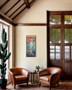 A tall, narrow abstract painting with vibrant pink, yellow, blue, and green tones hangs on a beige wall in a rustic room with two brown leather armchairs, a small side table, and dark wood double doors.