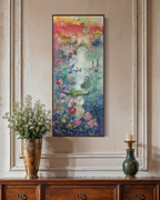 A tall, narrow colorful painting hangs on a beige wall above a wooden console table. The artwork features a whimsical fantasy scene with a white stag, flowers, birds, and a glowing sky in shades of blue, green, pink, and gold.