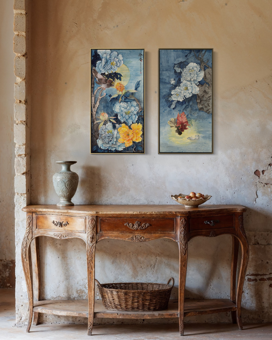 Two vertical framed floral paintings with blue, yellow, and white botanical tones hang on a textured beige plaster wall above an ornate wooden console table with a vase and bowl.