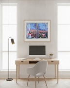 A framed art print depicting a window view of a pink flower field under a blue sky, displayed on a light gray wall above a wooden desk with a computer, chair, and floor lamp.