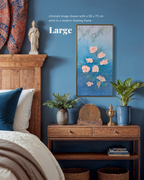 A framed wall art print featuring pink flowers on a blue background, shown hanging in a styled bedroom setting above a wooden console table with plants and decor.