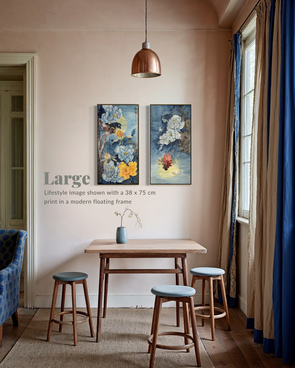 A lifestyle interior scene showing two vertical framed art prints hanging on a beige wall above a small wooden table and stools. The room has warm beige walls, blue curtains, a hanging copper-toned pendant light, and a rug.