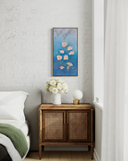 A vertical framed wall art print featuring pink flowers on a textured blue background, styled in a bedroom above a wooden nightstand with a vase of white flowers and a lamp.