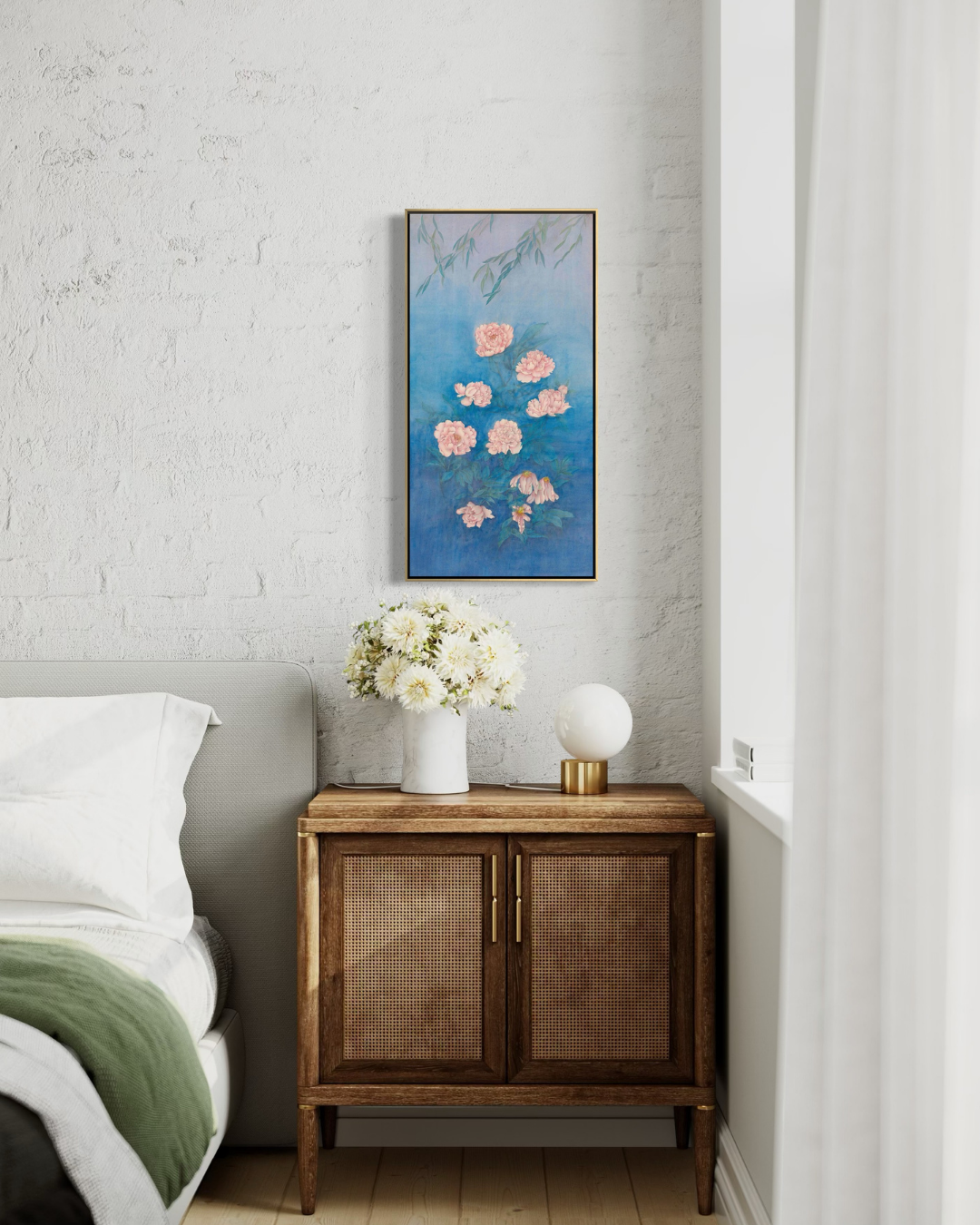 A vertical framed wall art print featuring pink flowers on a textured blue background, styled in a bedroom above a wooden nightstand with a vase of white flowers and a lamp.