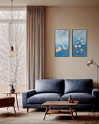 A styled living room scene featuring two vertical framed paintings with blue backgrounds and pink floral motifs, hung on a beige wall above a navy blue sofa.