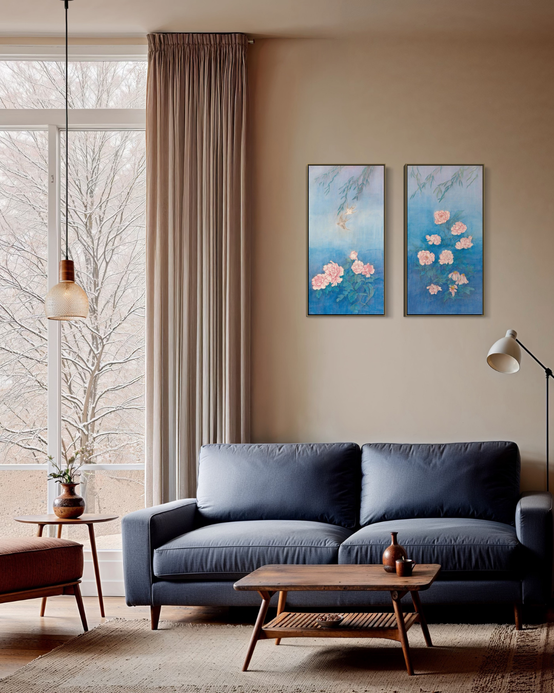 A styled living room scene featuring two vertical framed paintings with blue backgrounds and pink floral motifs, hung on a beige wall above a navy blue sofa.