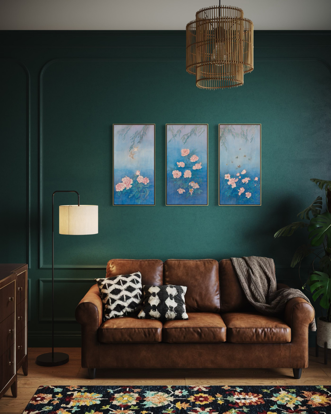 A styled living room featuring a dark green wall with three vertical framed paintings of pink cherry blossoms, a brown leather sofa with patterned pillows, a black floor lamp, and a woven pendant light.