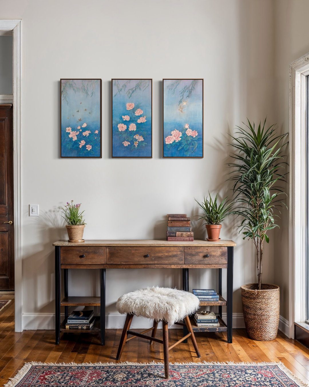 A styled interior scene showing a three-panel framed floral wall art set hanging above a wooden console table with a white faux-fur stool. The artwork features soft blue watercolor backgrounds with pink and peach blossoms.